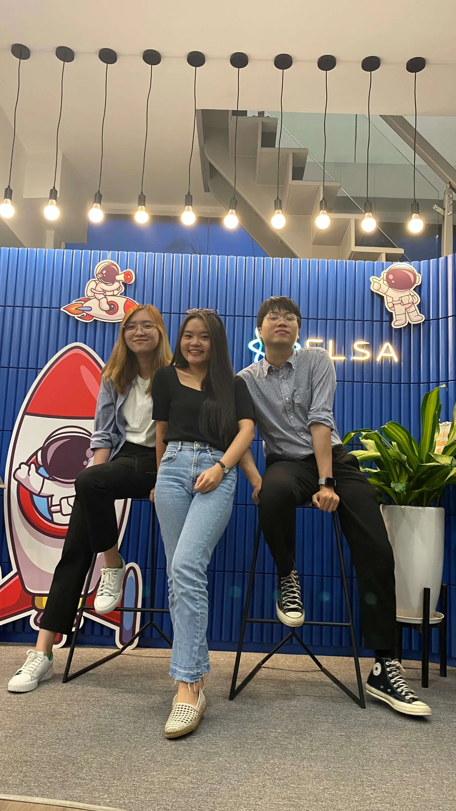 Three colleagues posing on bar stools in front of a blue office wall with astronaut and rocket cut-outs.