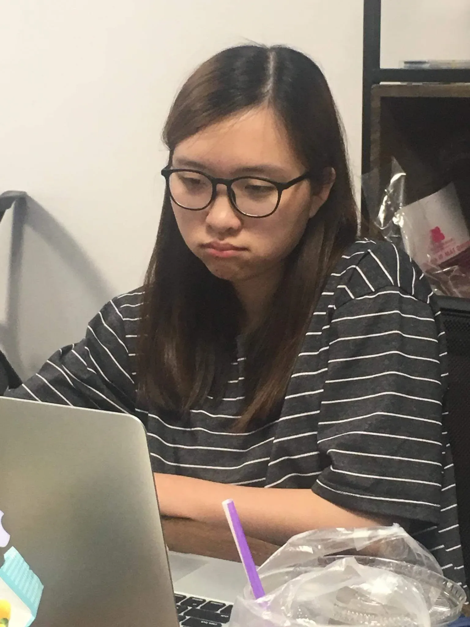 A woman in glasses and a striped T-shirt frowning at her laptop, a bubble tea beside it.