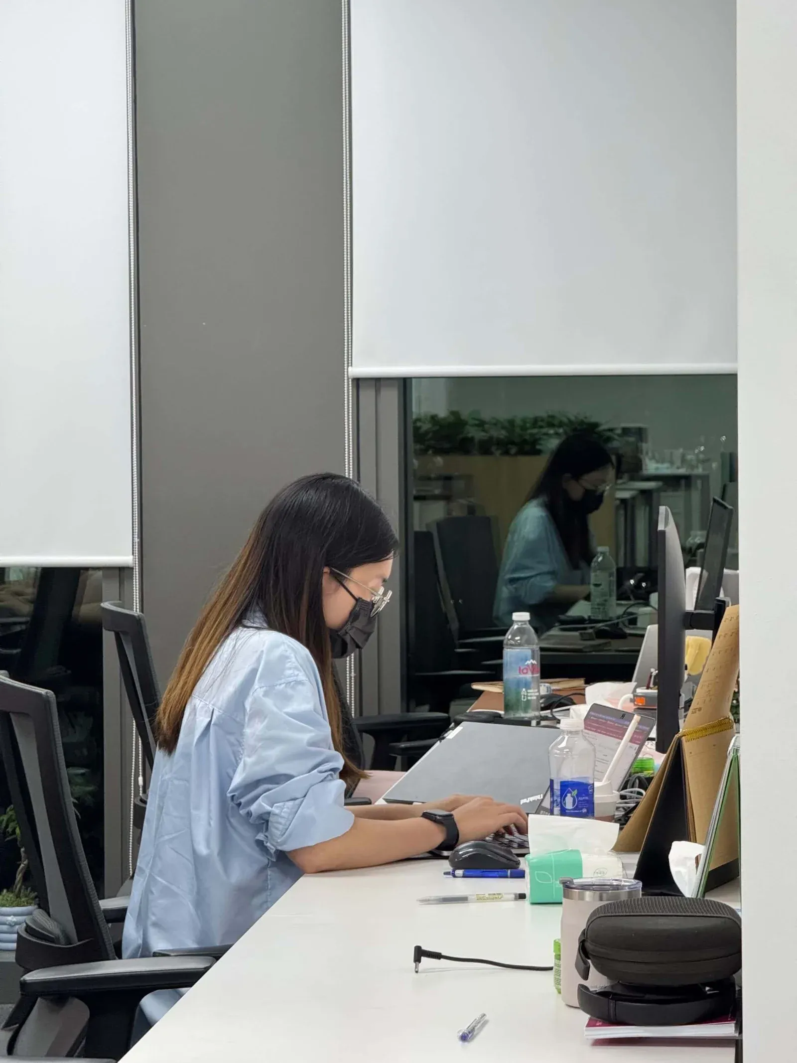 A woman in a mask and glasses typing at a desk in an open-plan office, her reflection in the glass behind her.