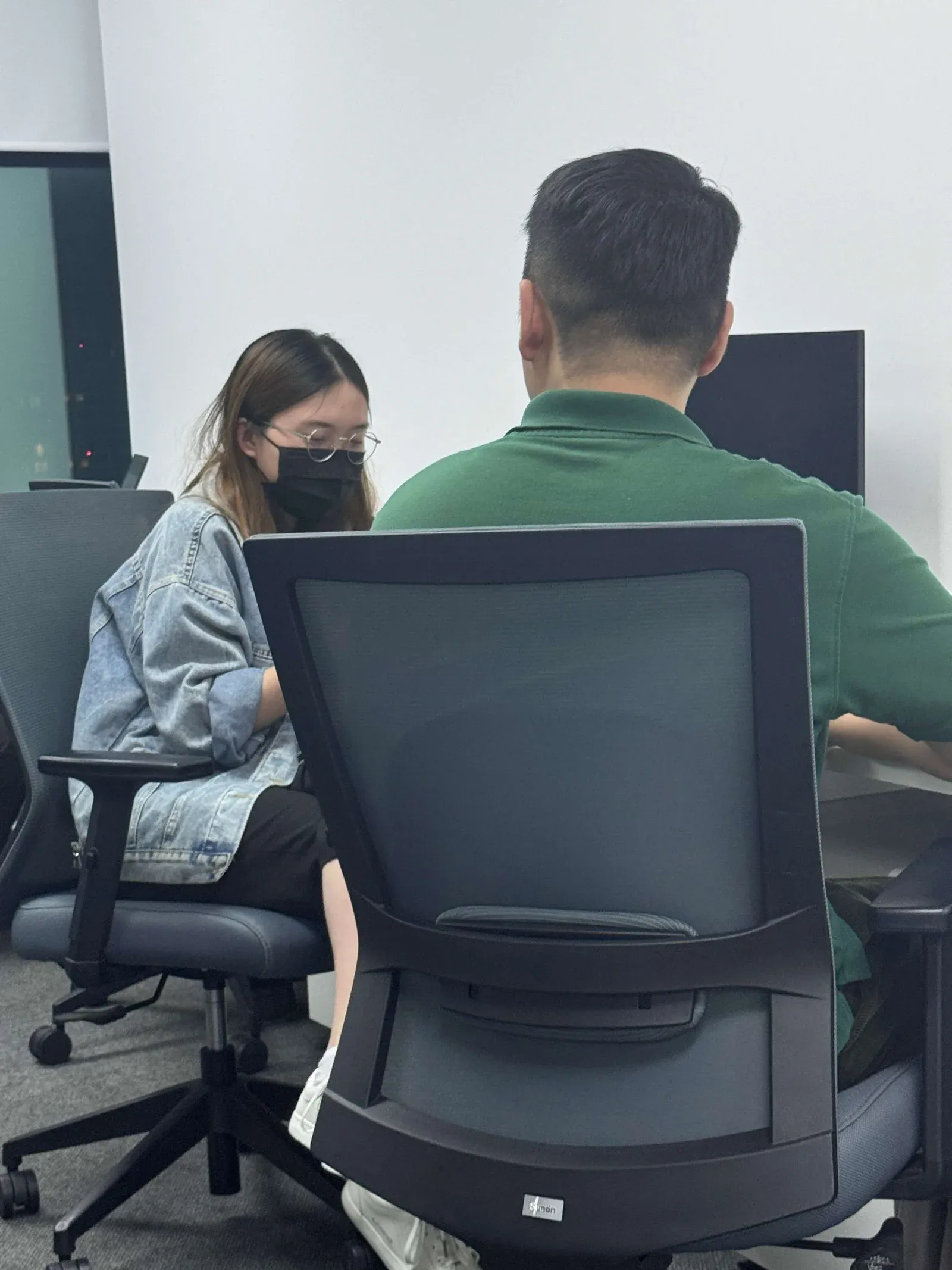 Two colleagues working side by side, one in a denim jacket and mask, the other seen from behind in a green polo.