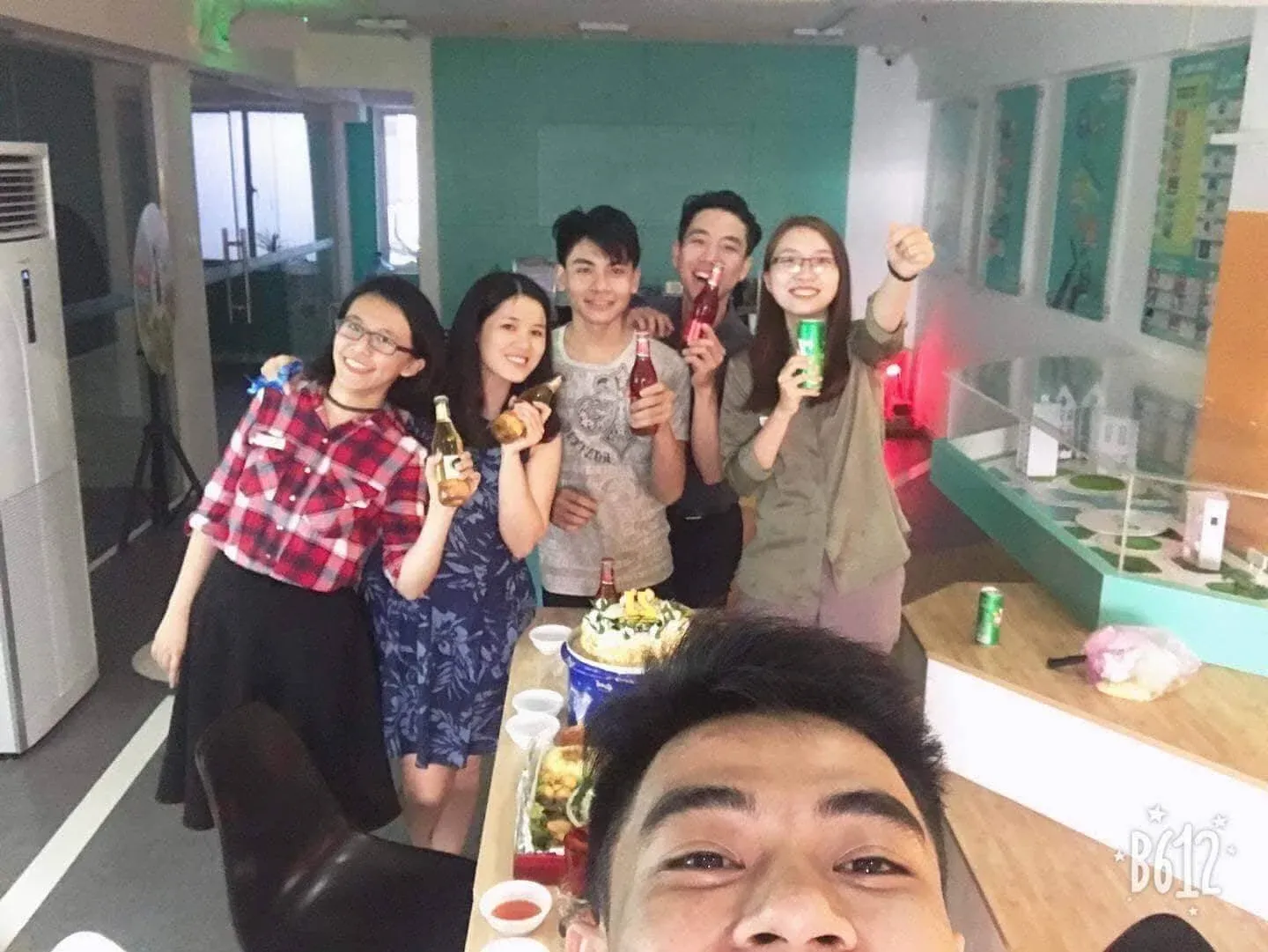 Colleagues holding bottles and cans around a table with a cake, one taking a selfie in the foreground.