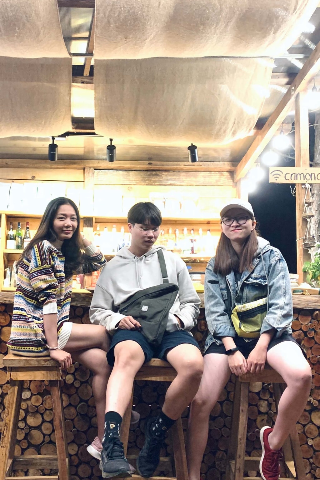 Three friends sit side by side on a wooden bench in a cozy café with warm lights and wooden beams.