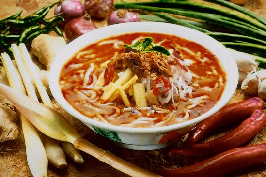 A bowl of tangy, spicy noodle soup in a red-orange broth, topped with fresh herbs and chili, with vegetables beside it.