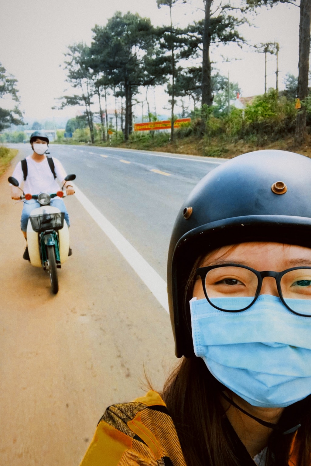 A selfie of a person in a helmet and face mask on a country road, with another rider and green hills behind.