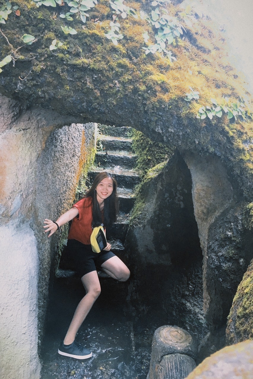 A woman in a red top climbs through a narrow gap in a mossy rock formation.