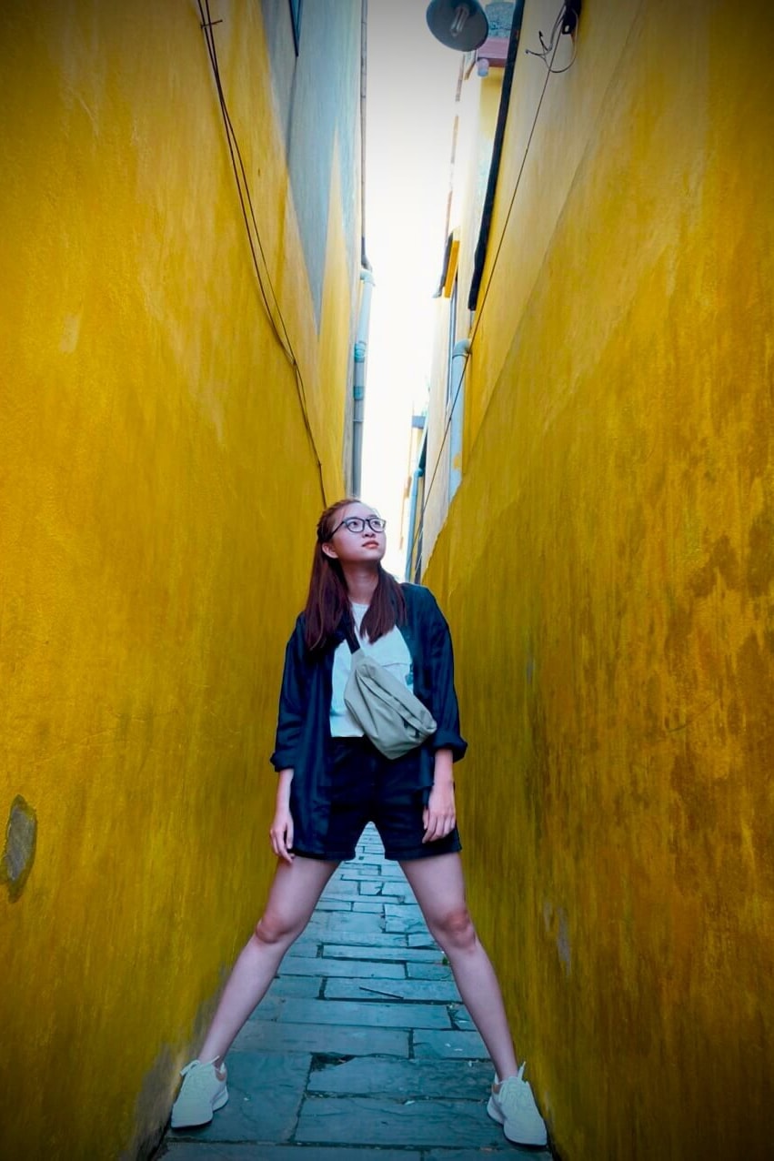 A woman in dark clothes stands in a narrow, yellow-walled alley in Hoi An’s old town.