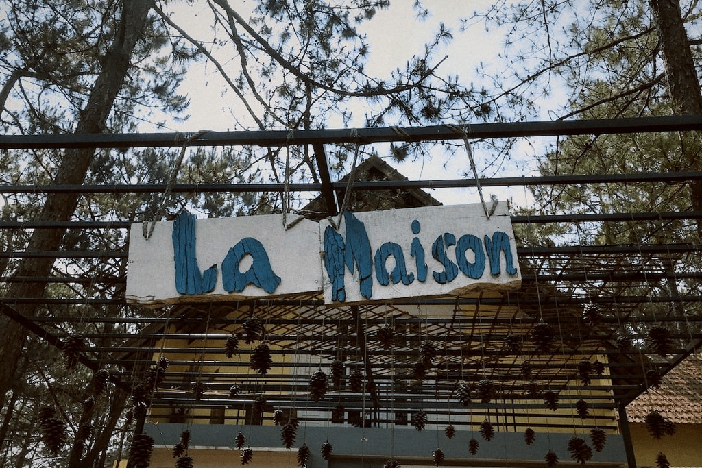 A hand-painted “La Maison” sign hangs among tree branches against a blue sky.