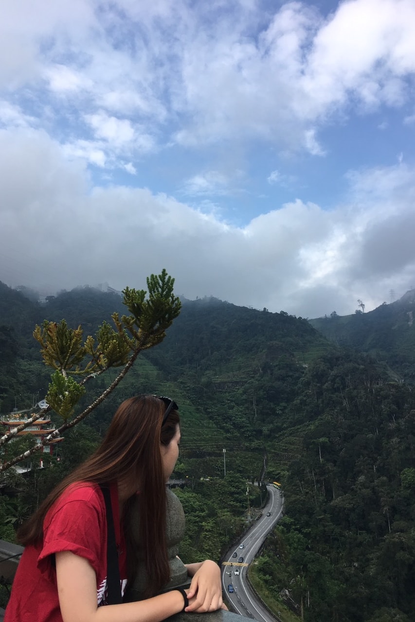 A woman in a red top looks out over misty green mountains under a cloudy sky.
