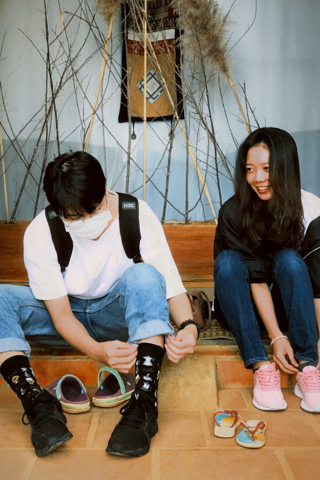 Two friends sit on a low step, bending down to tie their shoes; one wears a backpack and a face mask.