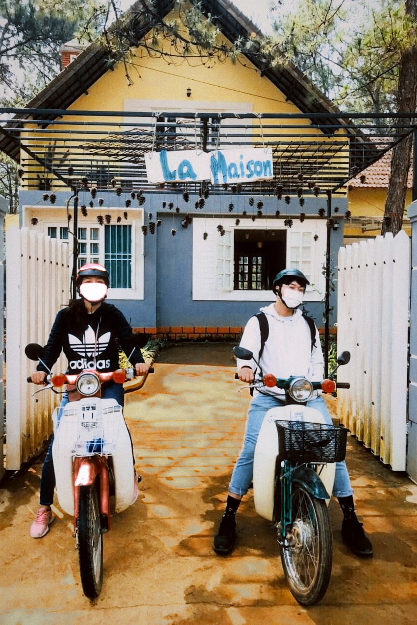 Two people on motorbikes in front of a small yellow cottage with a white fence.