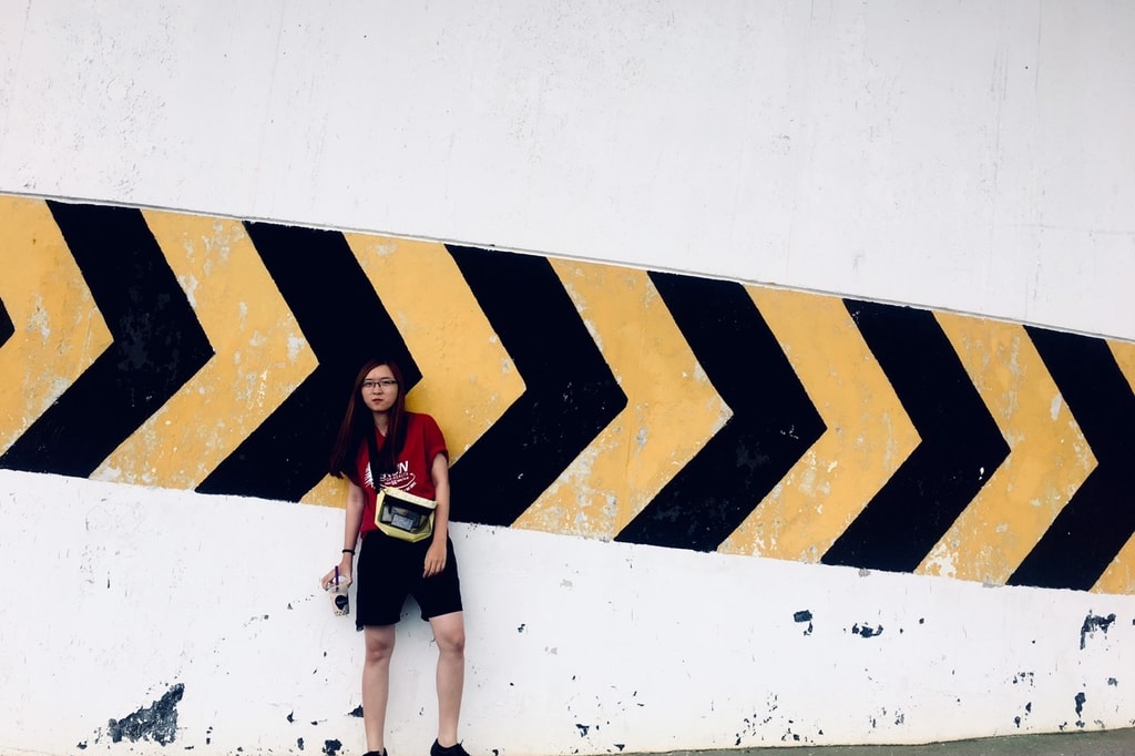 A woman in a red top and shorts stands against a white wall with a bold yellow and black arrow pattern.