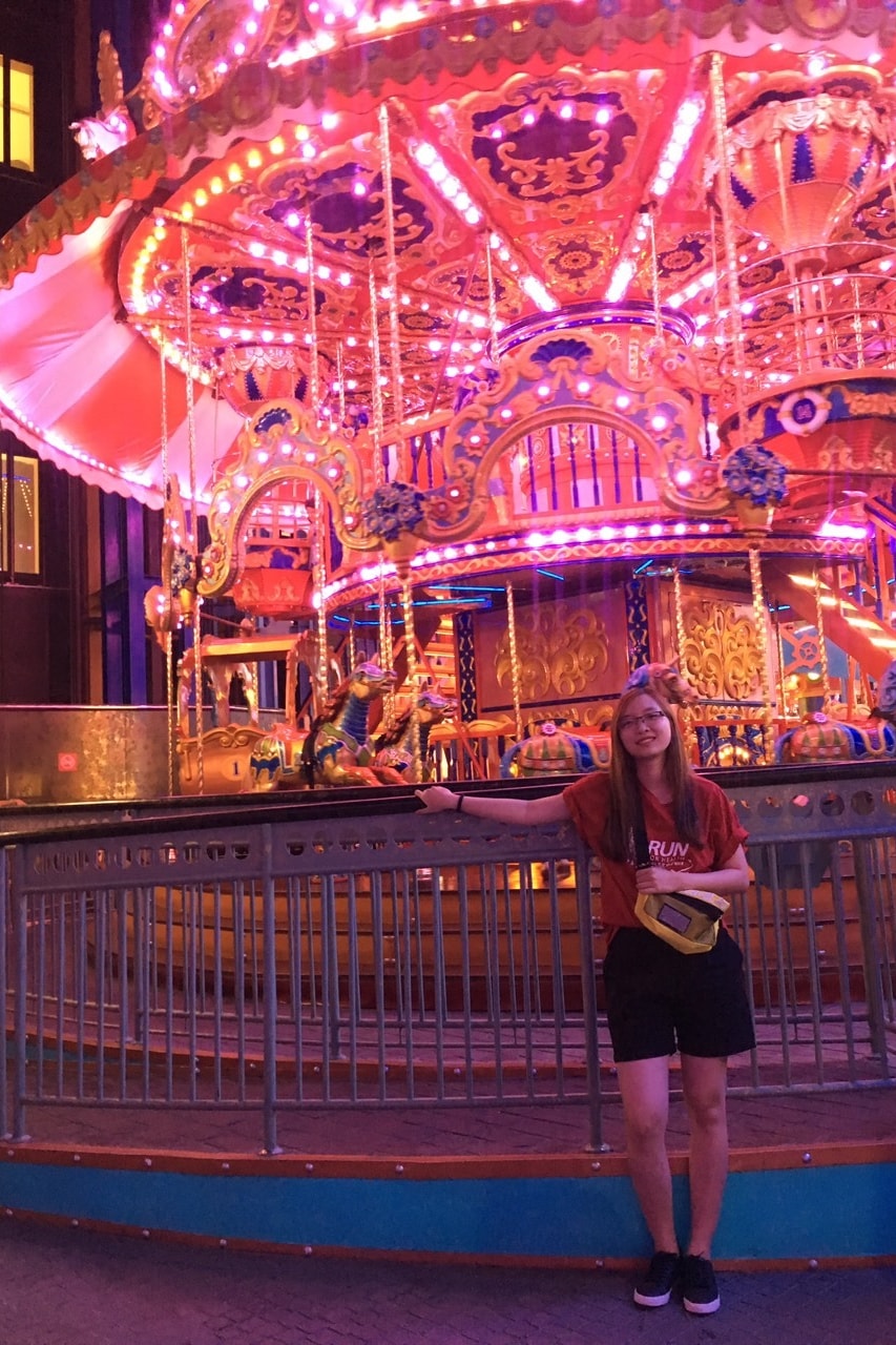 A woman stands in front of a carousel lit up in pink and red neon at night.